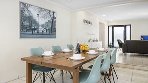 A dining area with a wooden table set for six, light blue chairs, and a cityscape painting on the wall. - Lovely 3br2ba w Wifi Parking in Central Location (Paradera)