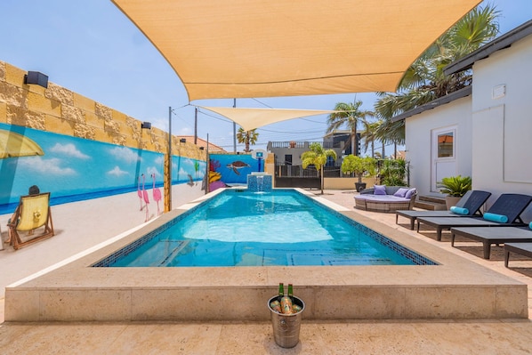 A swimming pool with lounge chairs and a shaded area, surrounded by a mural and palm trees.