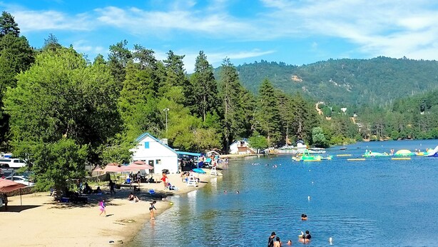 A lakeside beach with people swimming and sunbathing, surrounded by dense forest and hills.