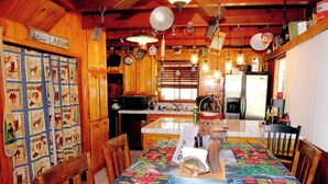 A wooden cabin interior with a dining table covered in a bear-themed quilt, wooden cabinets, and a kitchen area with a refrigerator and microwave.