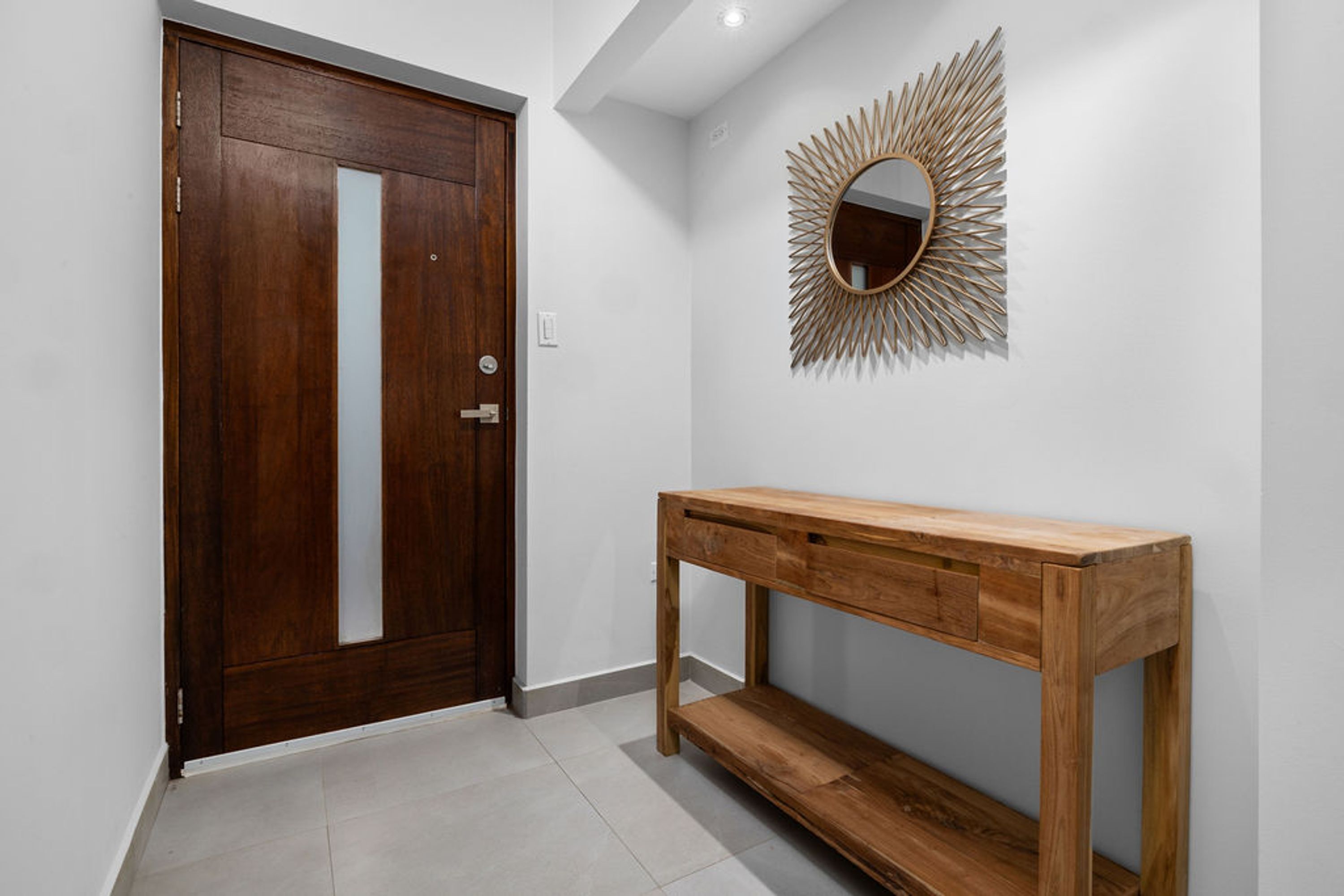 Un pasillo con una puerta de madera, una consola de madera y un espejo radiante en la pared.