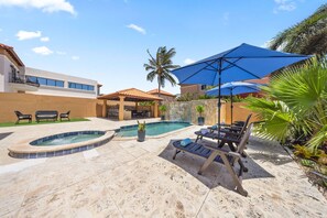 Ein Swimmingpool mit Liegestühlen und Sonnenschirmen, umgeben von einer Steinterrasse und Palmen.