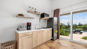 A modern kitchen with wooden cabinets, a microwave, a coffee maker, and a refrigerator. There are shelves above the counter holding jars and glasses. A sliding glass door leads to an outdoor area with a pool and patio furniture. - Tropical Getaway Near Arikok National Park Pool (Santa Cruz)