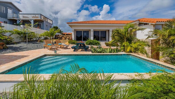 A swimming pool with lounge chairs and a patio area, surrounded by tropical plants and a two-story house with a red-tiled roof. - Tropical Getaway Near Arikok National Park Pool (Santa Cruz)