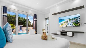 Quarto moderno com cama grande, TV fixada na parede e vista para a piscina e praia através da porta de vidro.