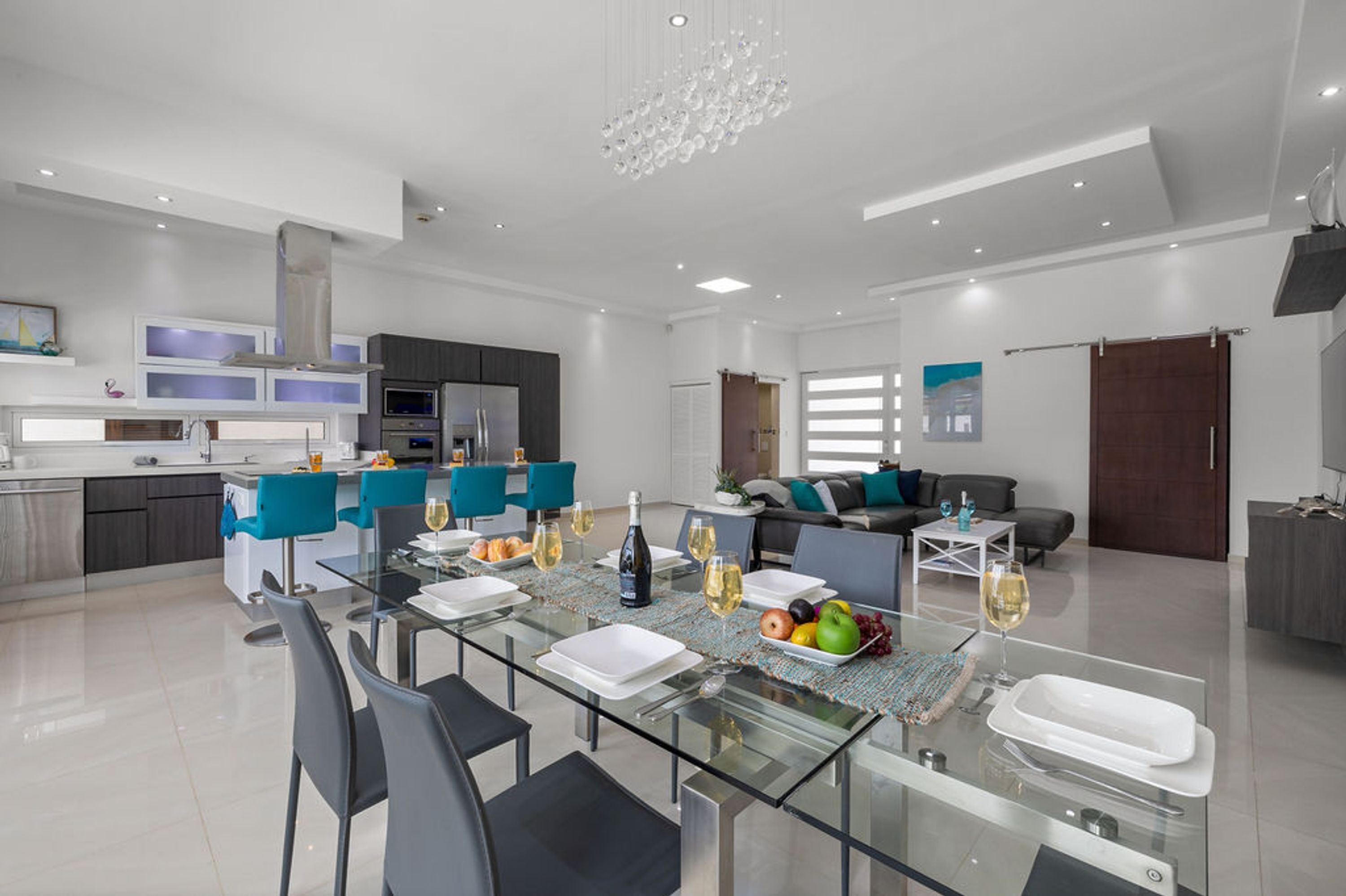 Una cuina i zona de menjador modernes, amb una taula de vidre preparada per a dinar, una sala d'estar amb sofà i taula de centre, i un gran candelabrum.