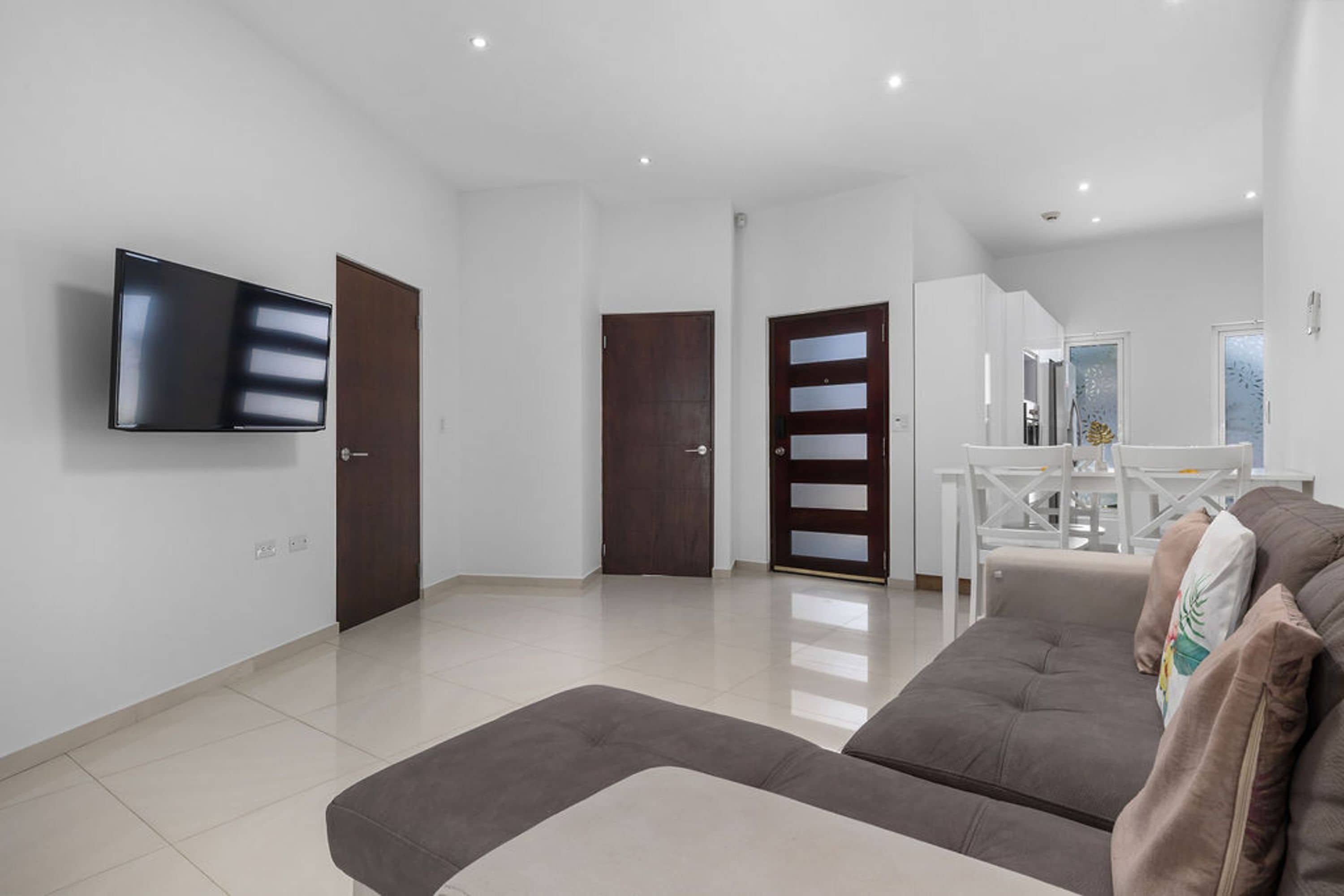 Una sala de estar moderna con un televisor de pantalla plana montado en la pared, un sofá y un comedor con una mesa y sillas blancas.