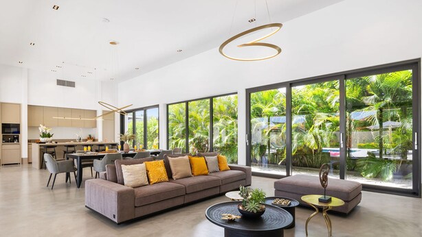 Ein modernes Wohnzimmer mit einer Couch, einem Couchtisch und einem Hocker. Es gibt eine Essecke mit Stühlen und einem Tisch. Große Fenster bieten Blick ins Grüne.