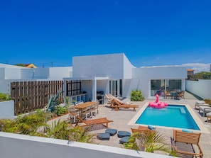 Un área de piscina moderna al aire libre con terraza de madera, sillas de descanso y un flamenco inflable rosa.