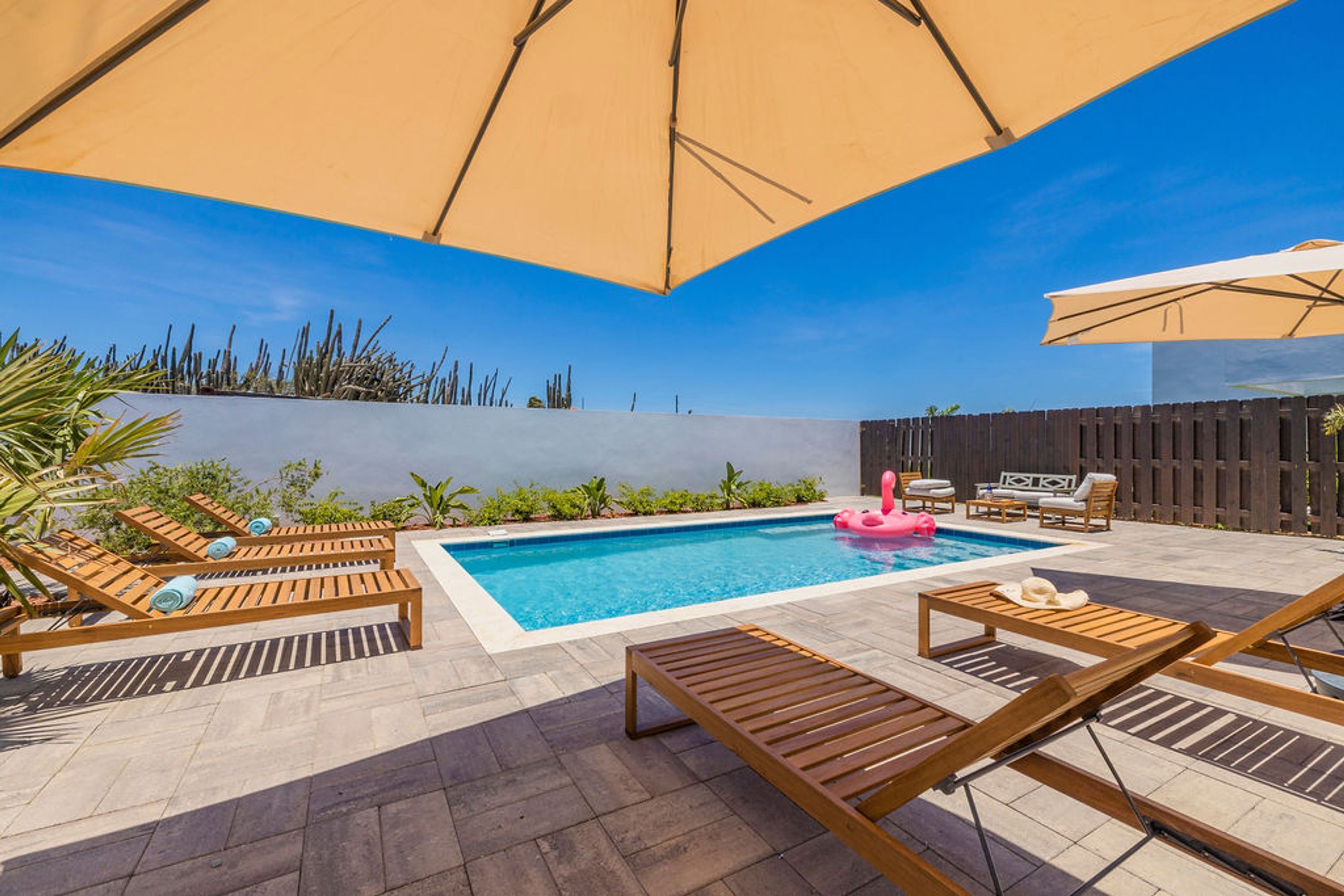 A pool area with wooden loungers, a pink flamingo float, and umbrellas.