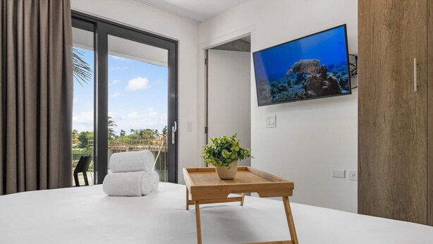 A modern hotel room with a large bed, a wooden side table with a plant, a TV on the wall, and a view of the outdoors. - Serene Designer Gem Walk to Beach Strip Pool (Noord)