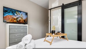 A modern bedroom with a TV, a wooden side table with a cup and berries, and rolled towels.