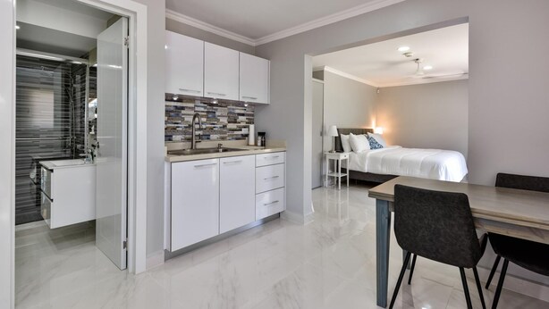 Una cucina moderna con mobili bianchi e zoccolo piastrellato, una zona pranzo con tavolo e sedie in legno, e una camera da letto con un letto e un comodino.