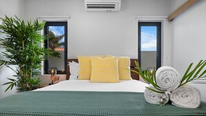 Une chambre d’hôtel avec un lit, une couvre-lit verte, deux fenêtres, une plante en pot et un climatiseur fixé au mur.
