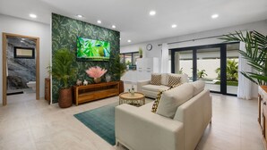 Un salon moderne avec un grand téléviseur à écran plat, un canapé, une table basse et un meuble en bois. Il y a une grande fenêtre avec des rideaux, une plante et une vue sur l’extérieur.