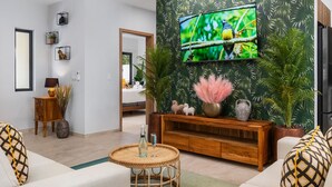 Un salon avec un téléviseur fixé au mur, orné d’un motif de feuilles tropicales, une console en bois, une table basse ronde en osier et un canapé agrémenté de coussins à motifs.