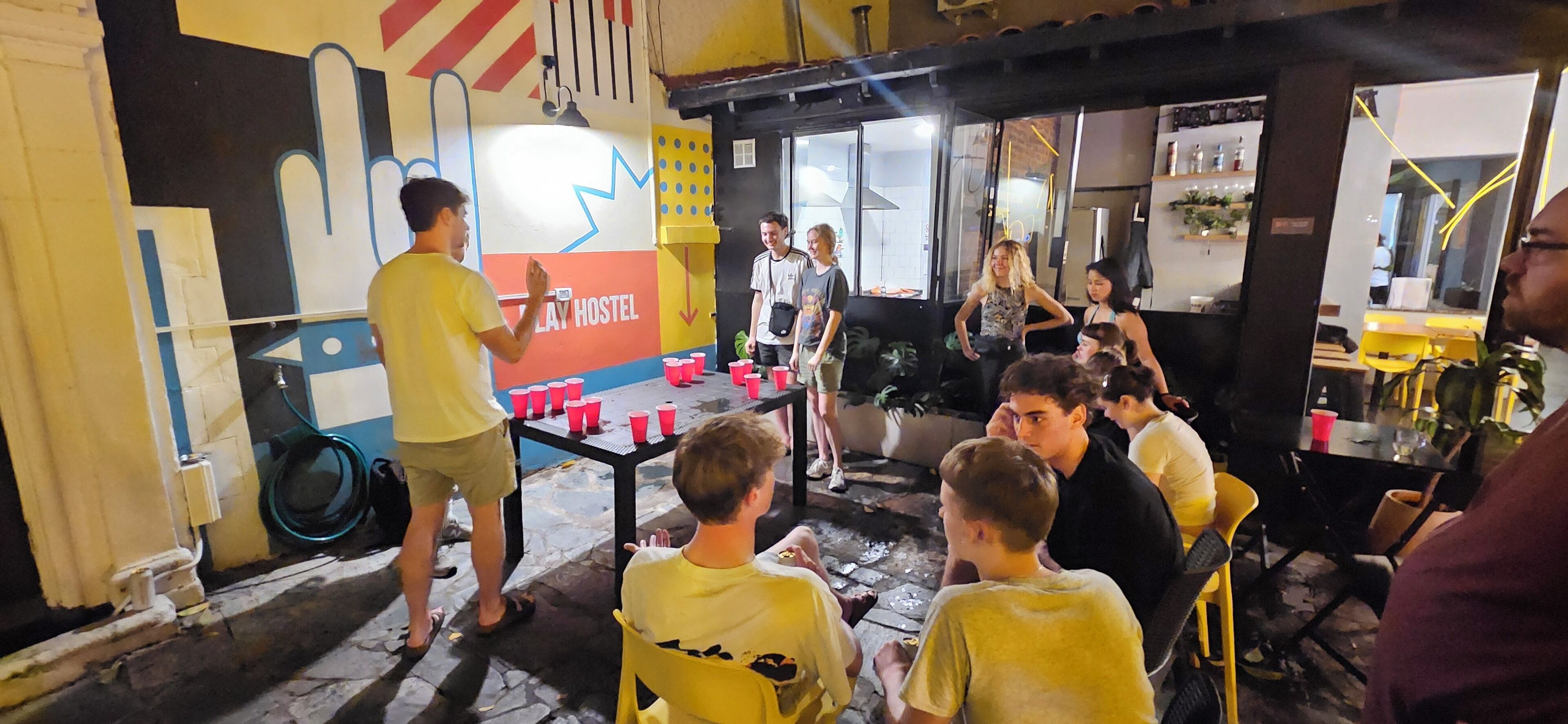 Interior de un hostal con zona de juegos, una mesa con vasos rojos y un mural en la pared.