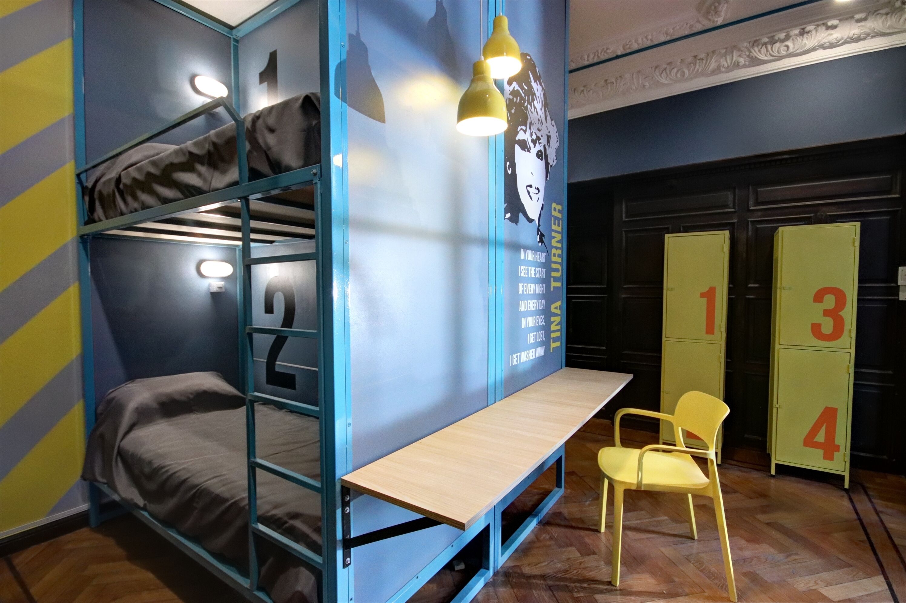 Una cama individual superpuesta con escritorio y silla, una pared con un cartel, y taquillas numeradas del 1 al 4.