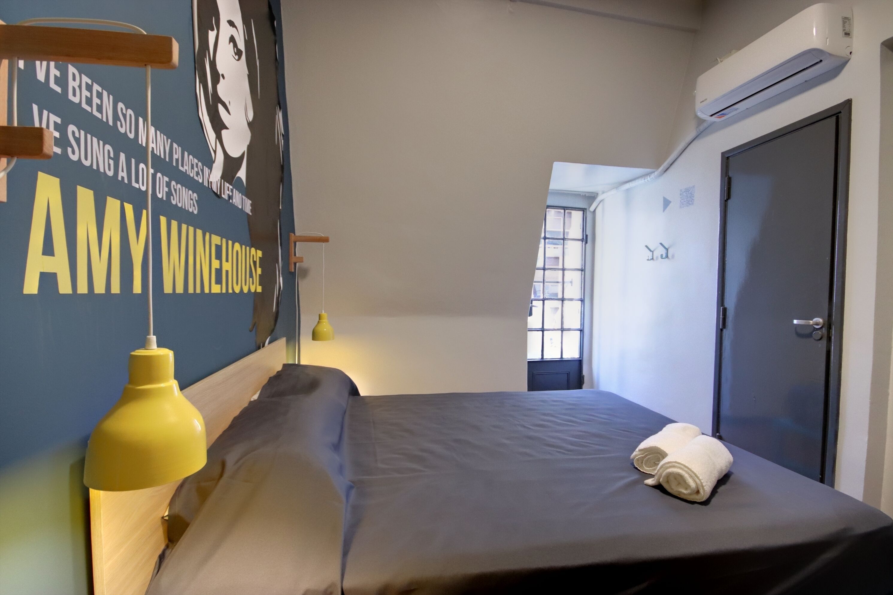 A hotel room with a bed, a wall poster, a yellow pendant light, and an air conditioning unit.