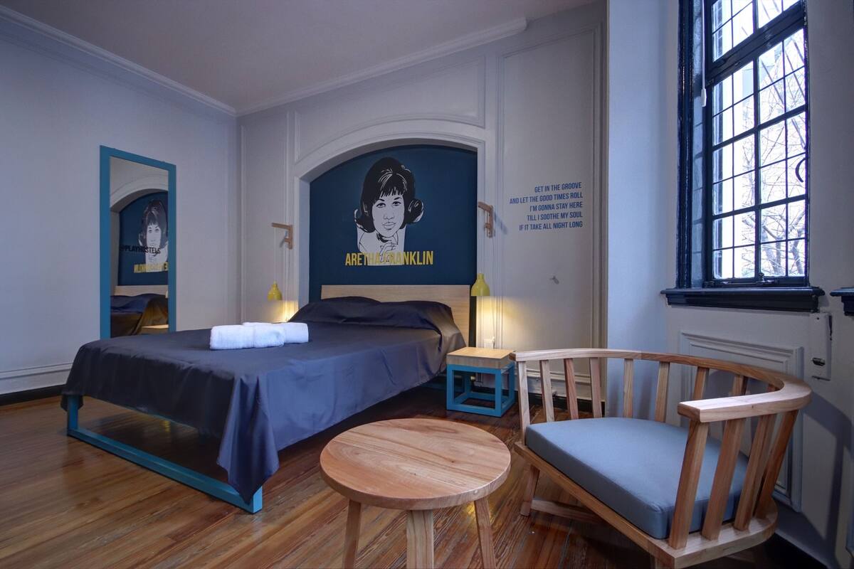 Hotellihuone, jossa on sänky, pyöreä puinen pöytä, puinen tuoli, peili ja ikkuna.