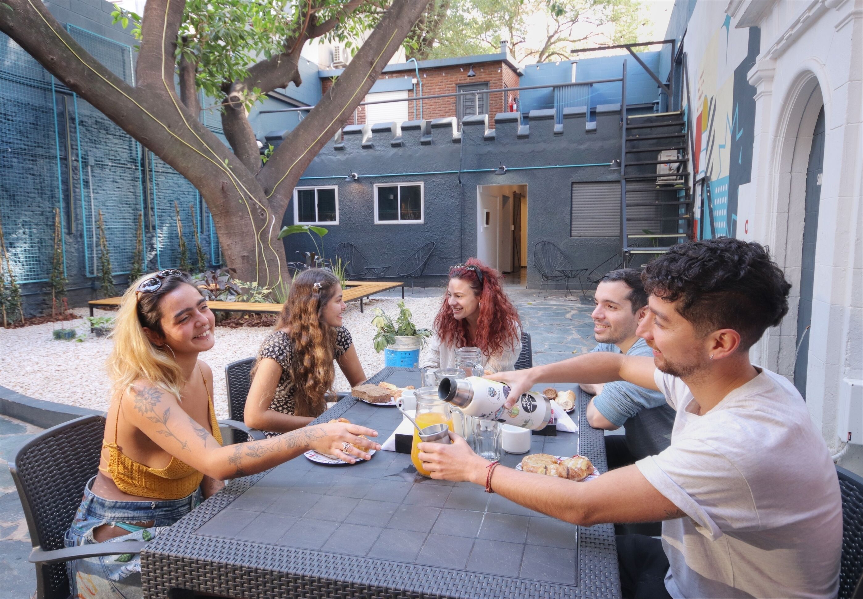 A group of people seated around a table outdoors with a tree and a building in the background.