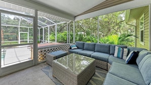 A covered outdoor seating area with a sectional sofa and a glass-top coffee table, surrounded by large windows offering views of lush greenery. - Garden House with Pool & Spa (Bradenton)