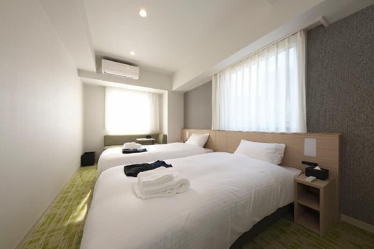 A hotel room with two beds, a bedside table, a lamp, a window with curtains, and an air conditioning unit.