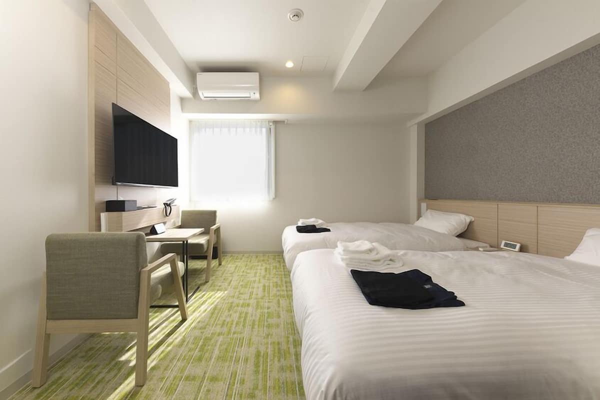 A hotel room with two beds, a desk with a chair, a flat-screen TV, and an air conditioning unit.