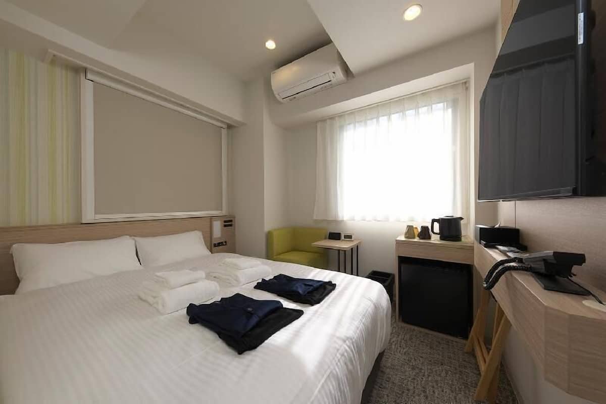 A hotel room with a bed, a desk, a chair, a television, and an air conditioning unit.