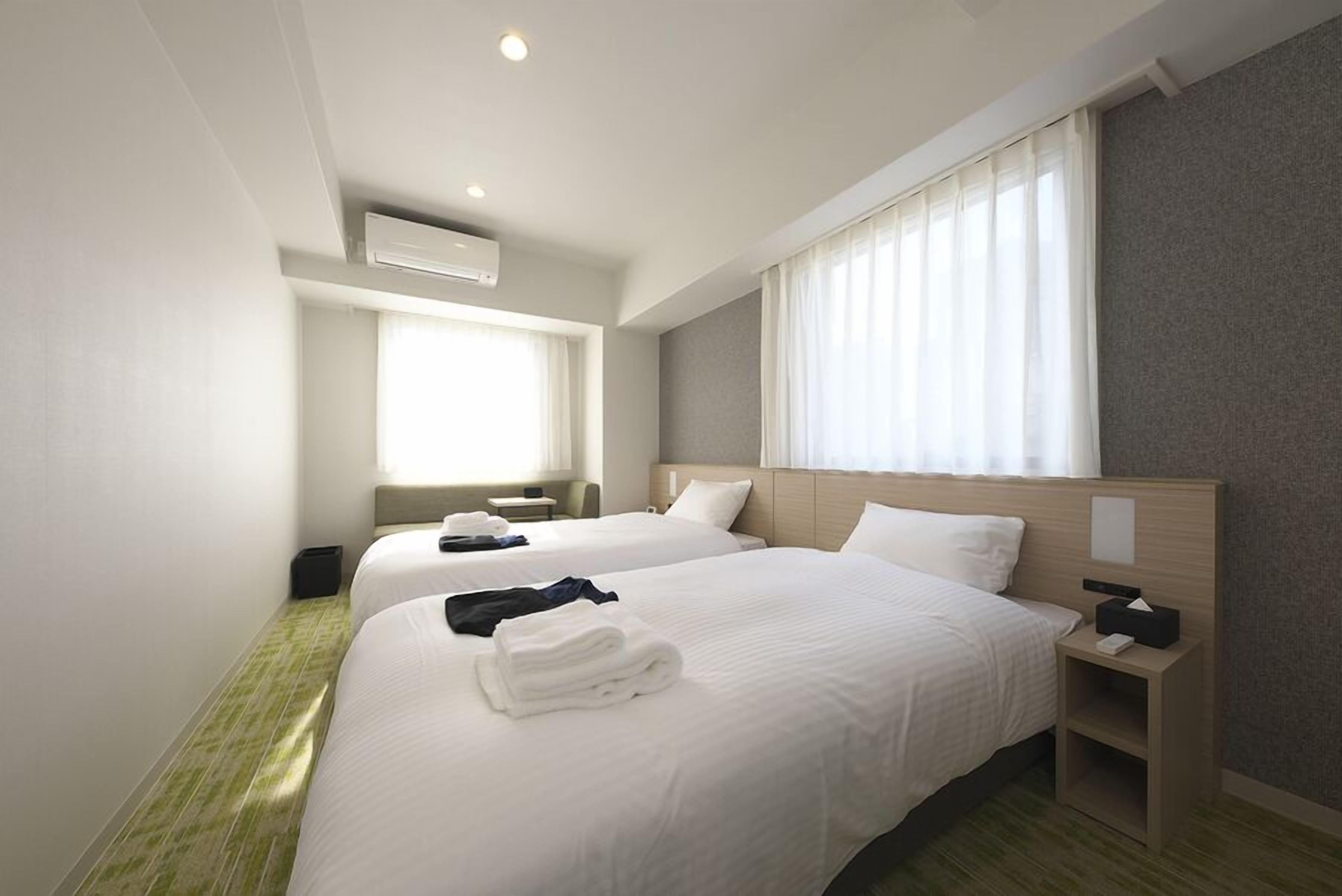 Een hotelkamer met twee bedden, een nachtkastje met een lamp, een raam met gordijnen en een airconditioning.