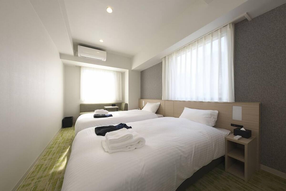 A hotel room with two beds, a bedside table with a lamp, a window with curtains, and an air conditioning unit.