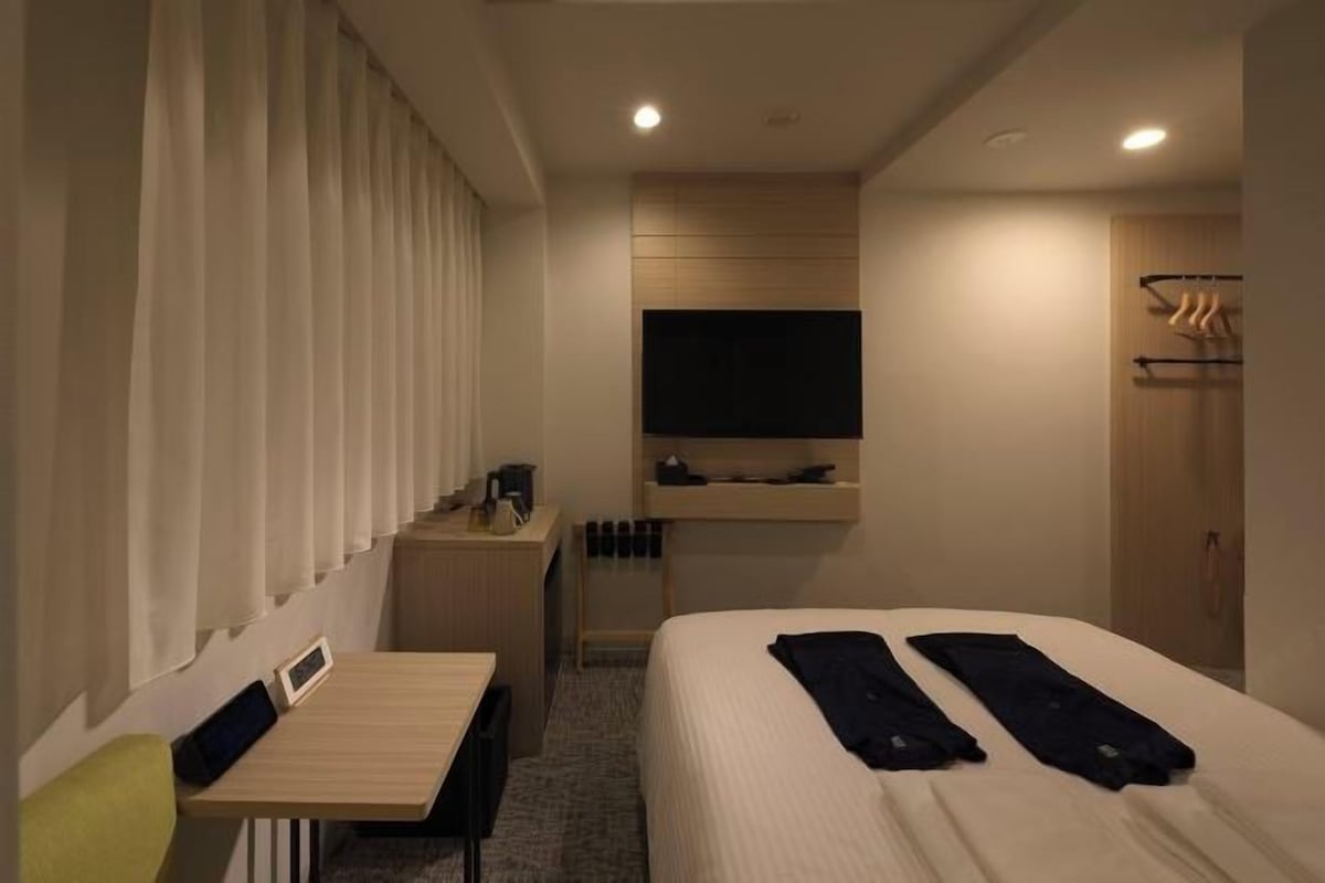 A hotel room with a bed, a desk, a chair, a TV mounted on the wall, and a coat rack.