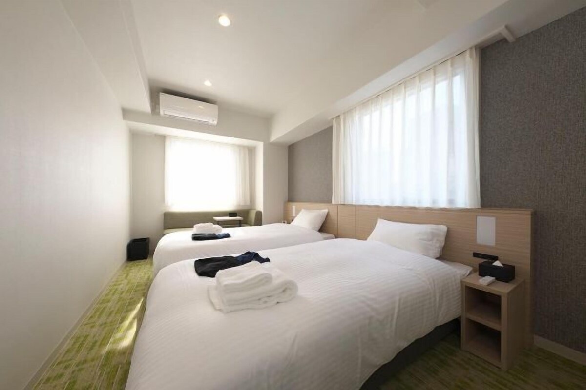 Een hotelkamer met twee bedden, een nachtkastje met een lamp, een raam met gordijnen en een airconditioning.