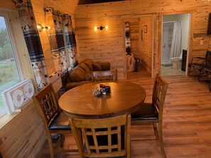 Een houten interieur met een ronde eettafel, stoelen en een bank. Er is een raam met gordijnen en een zichtbare badkamer.