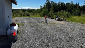 A gravel parking area with picnic tables and a building with a white exterior. - RV Site with Full Hookups (Sterling)