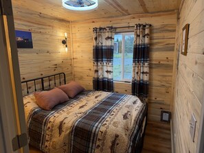 Et soveværelse i en træhytte med seng, puder, et vindue med gardiner og en lampe monteret på væggen.