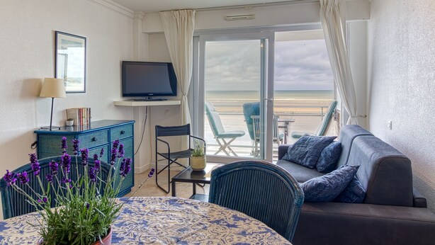 A room with a balcony, a sofa, a TV, a table with a plant, and a view of the beach. - Les Goélands - Vue mer à moins d'1 Km de la plage (Bray-Dunes)