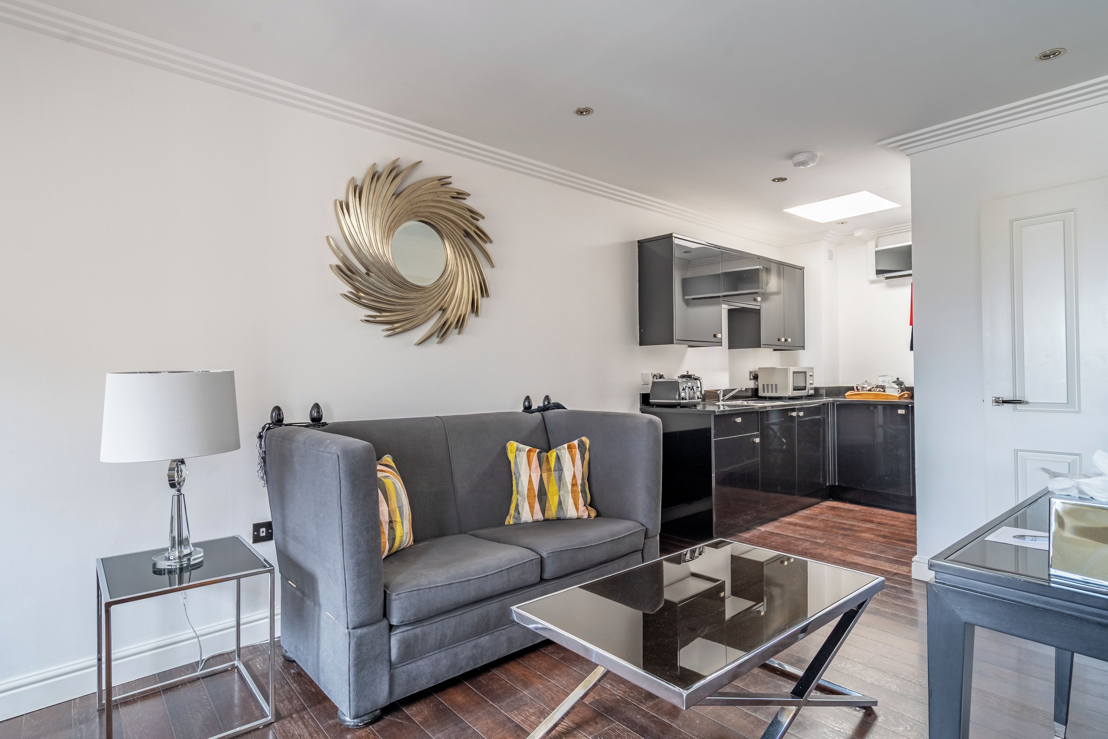 Uma sala de estar moderna com um sofá cinzento, uma mesa de centro de vidro e um espelho decorativo na parede.