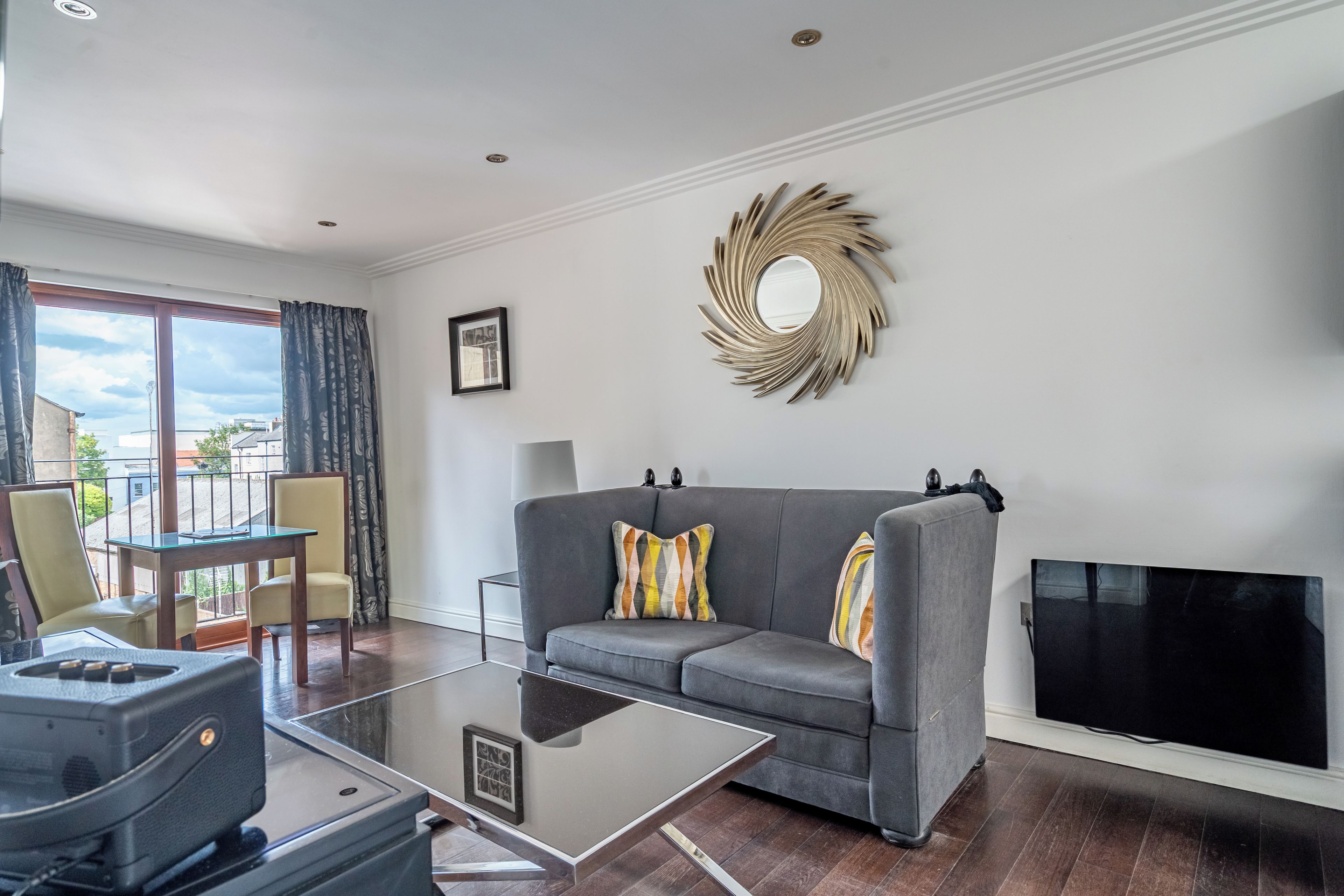 Una sala de estar moderna con un sofá gris, una mesa de centro de vidrio, un televisor de pantalla plana y un espejo decorativo en la pared.