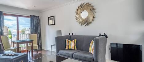 A modern living room with a grey sofa, a glass coffee table, a flat-screen TV, and a decorative mirror on the wall.