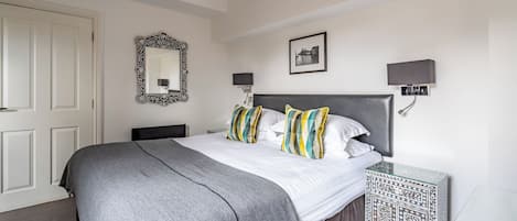 A neatly arranged bedroom with a large bed, a bedside table, a mirror, and a wall-mounted lamp.