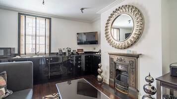 A modern living room with a fireplace, a large mirror, and a kitchen area with black cabinets and a flat-screen TV.