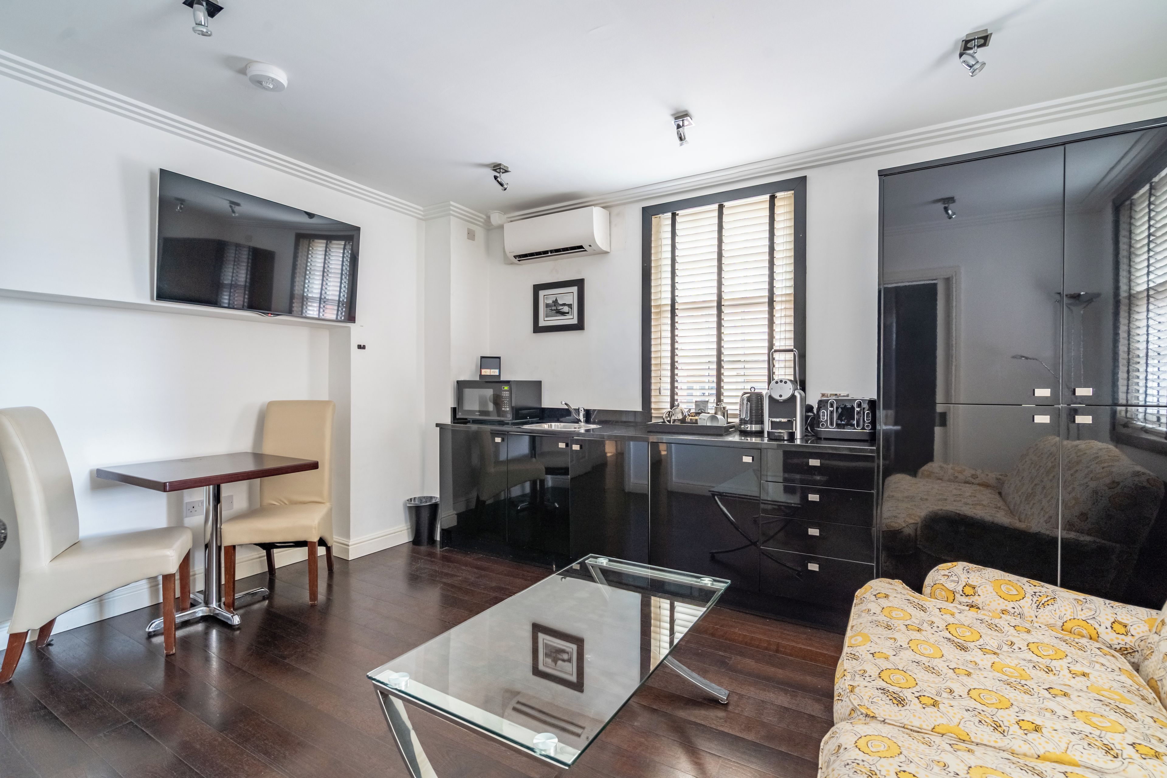 A modern living room with a glass coffee table, a sofa with a patterned cover, a dining area with a table and chairs, a kitchen with a black countertop, and a large mirror reflecting the room.