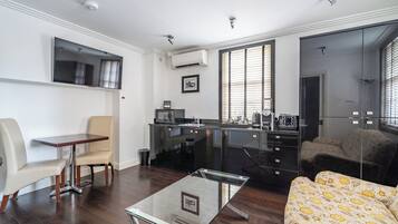 A modern living room with a glass coffee table, a sofa with a patterned cover, a dining area with a table and chairs, a kitchen with a black countertop, and a large mirror reflecting the room.