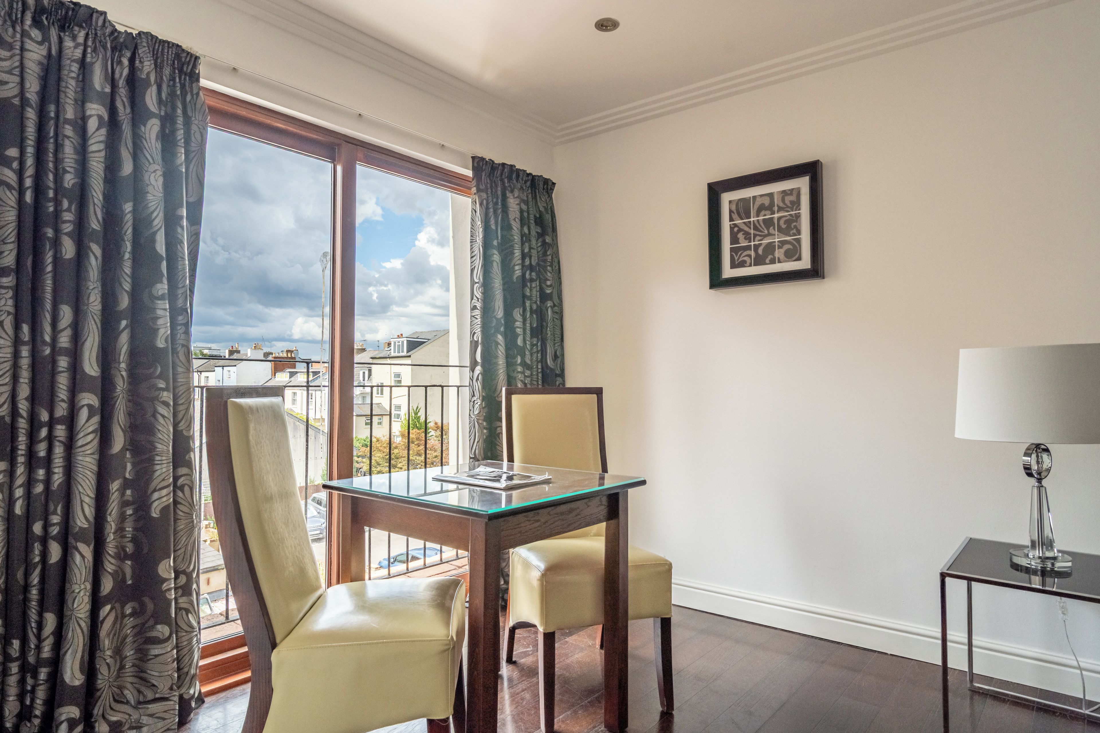 A room with a glass-top dining table, two chairs, a small side table with a lamp, and a framed picture on the wall.