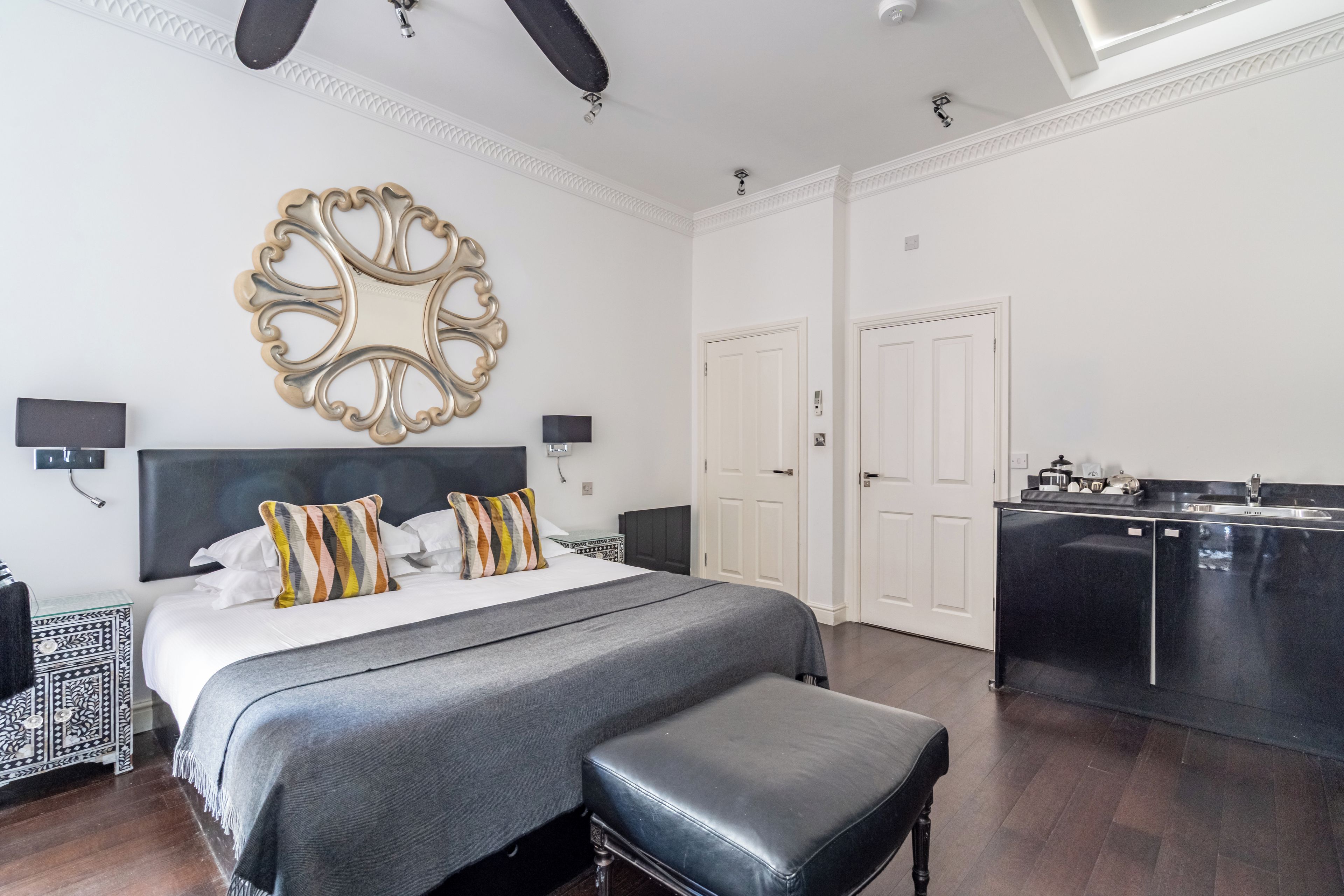 A modern bedroom with a large bed, bedside tables, a black bench, a decorative mirror, and a kitchen area.