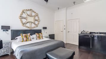 A modern bedroom with a large bed, bedside tables, a black bench, a decorative mirror, and a kitchen area.