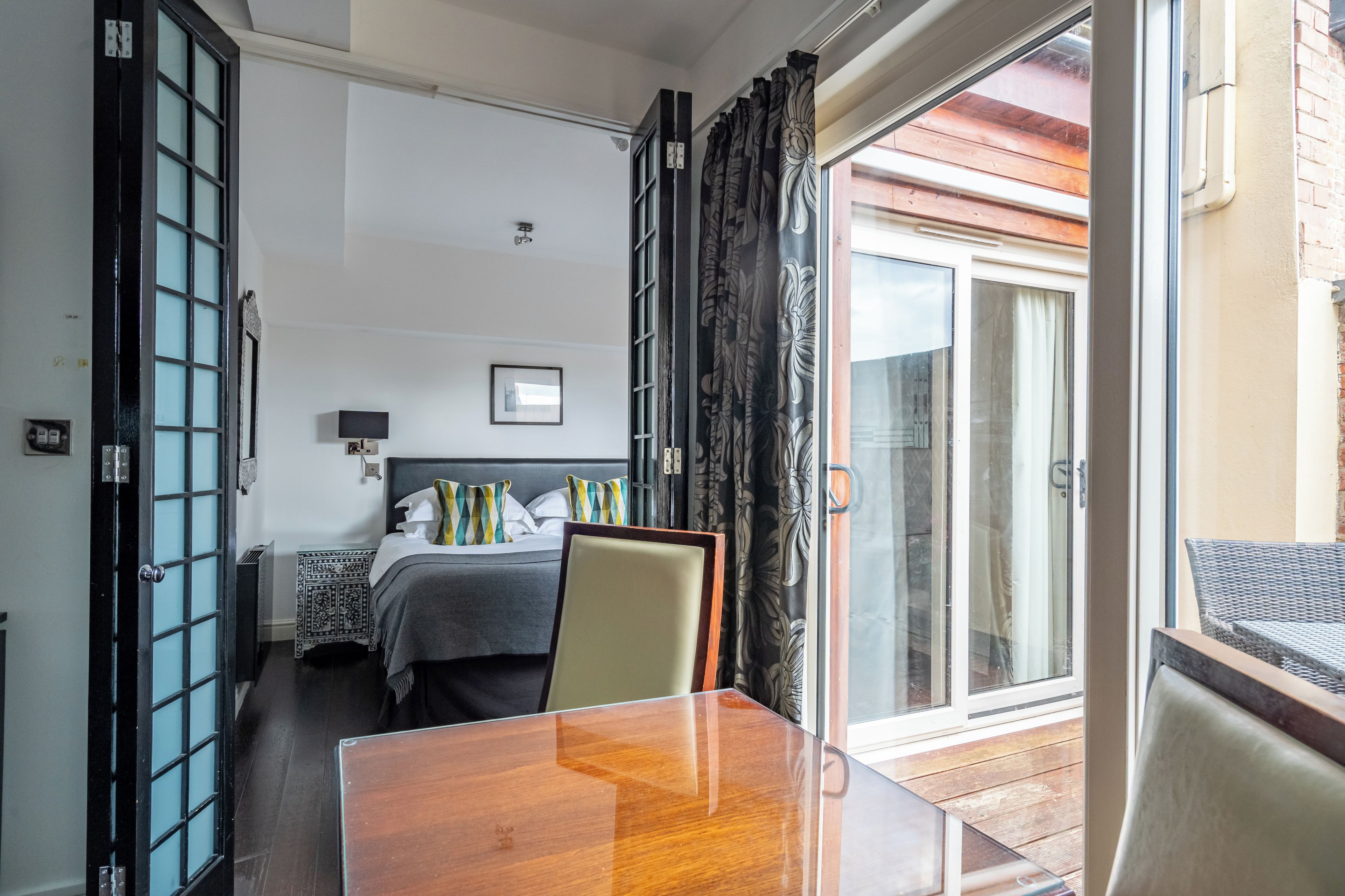 A hotel room with a wooden table, a bed with a grey headboard, a nightstand, and a view of the outside through a glass door.
