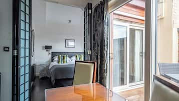 A hotel room with a wooden table, a bed with a grey headboard, a nightstand, and a view of the outside through a glass door.