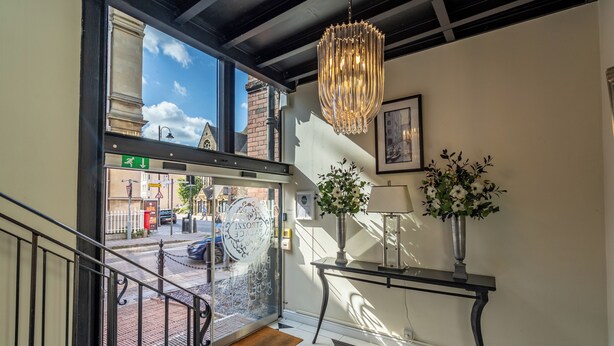 A modern hotel lobby with a glass door leading outside, a chandelier, a table with a vase of flowers, and a framed picture on the wall.